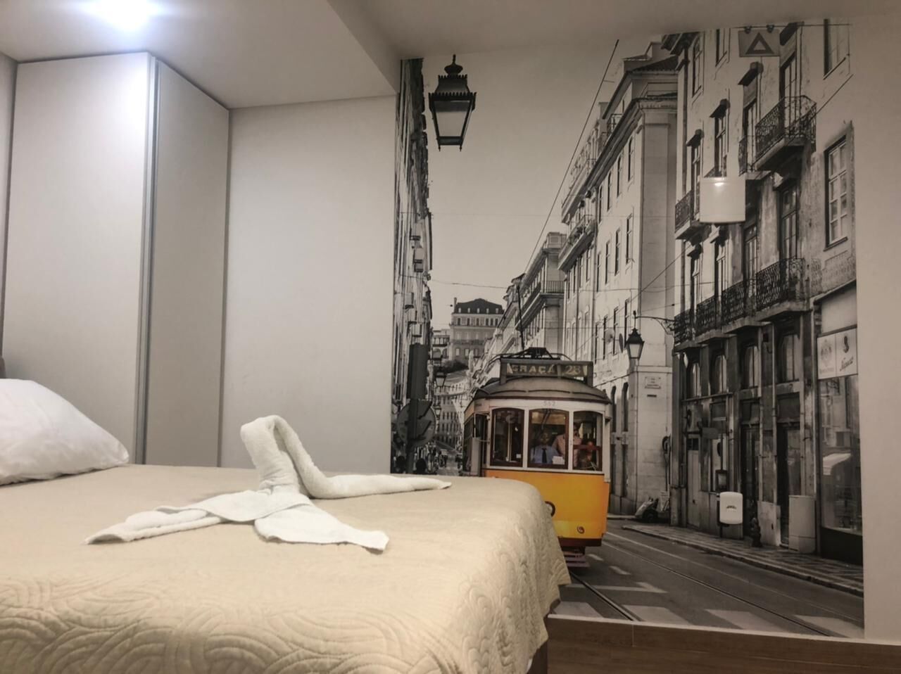 Une chambre d’hôtel avec un lit, une serviette blanche et une fresque murale représentant une scène de rue avec un tramway.