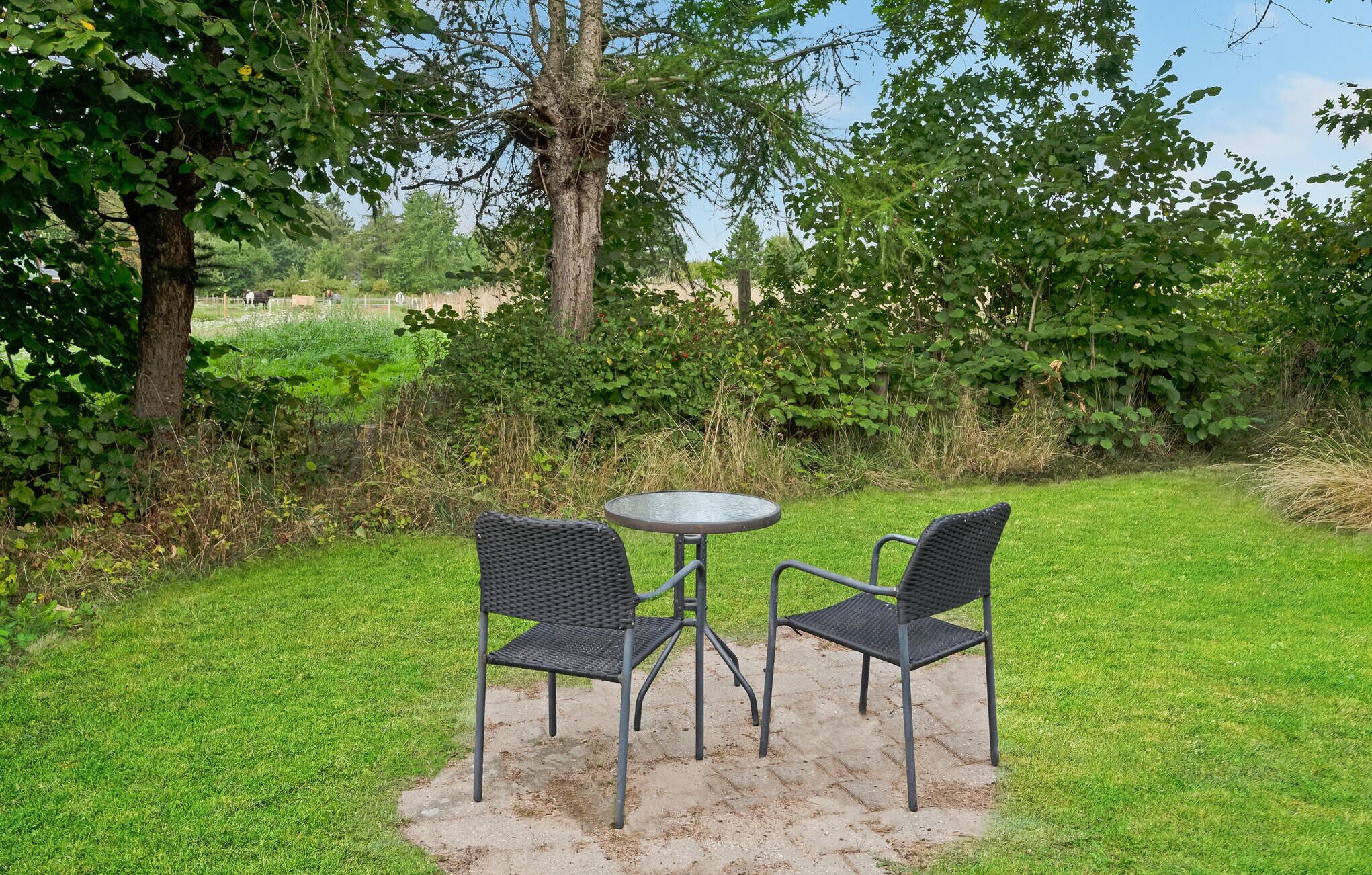Eine kleine Sitzgelegenheit im Freien mit zwei Stühlen und einem Tisch, umgeben von Grünpflanzen und Bäumen.