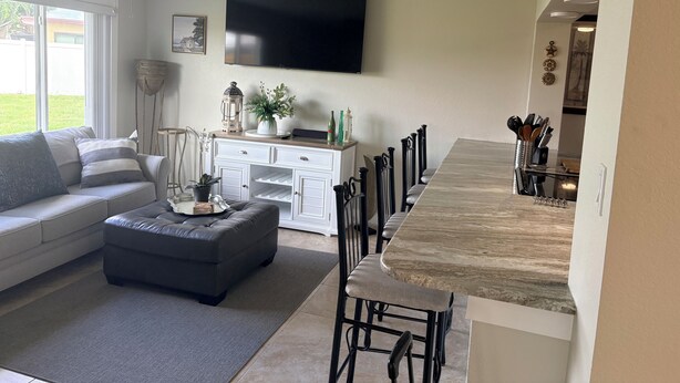 A modern living room with a grey sofa, a dark ottoman, a white cabinet, a flat-screen TV, and a dining area with a marble countertop and black chairs.