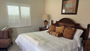 A neatly made bed with a white comforter and decorative pillows, a wooden nightstand with a lamp, a brown armchair, and a window with blinds.