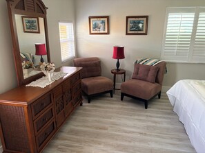 A bedroom with a wooden dresser, two chairs, a bed, and two windows with blinds.