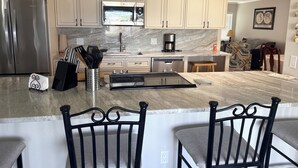 A kitchen with a stainless steel refrigerator, a microwave, a coffee maker, and a sink. There are two bar stools and a dining table.