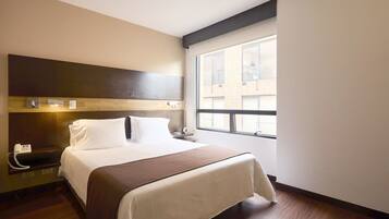 Habitación de hotel con cama, mesita de noche, lámpara y ventana con vistas a edificios.