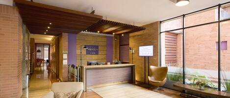 A hotel reception area with a reception desk, a waiting area with a sofa, and a large window offering a view of the outdoors.