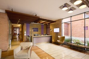 A hotel reception area with a reception desk, a waiting area with a sofa, and a large window offering a view of the outdoors. - Mercure bh Retiro (Bogotá)