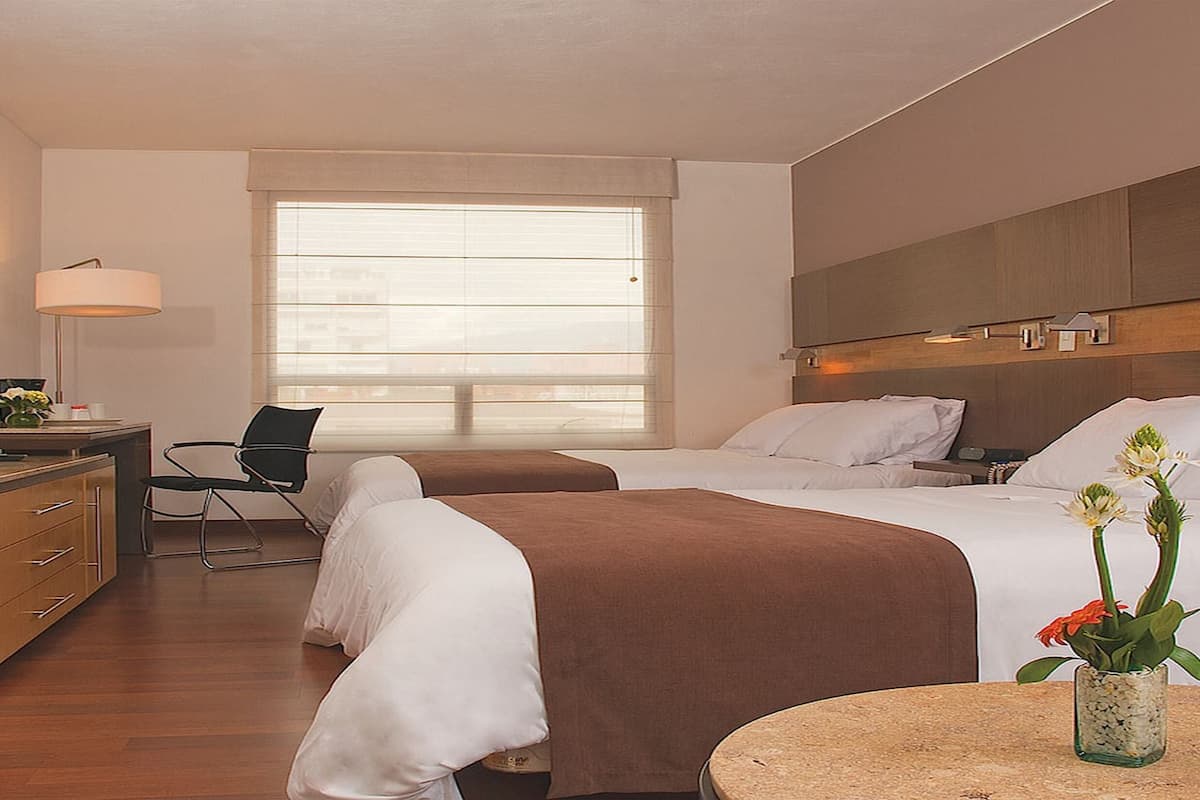 Quarto de hotel com duas camas, uma mesa, uma cadeira, uma televisão e uma janela com persianas.