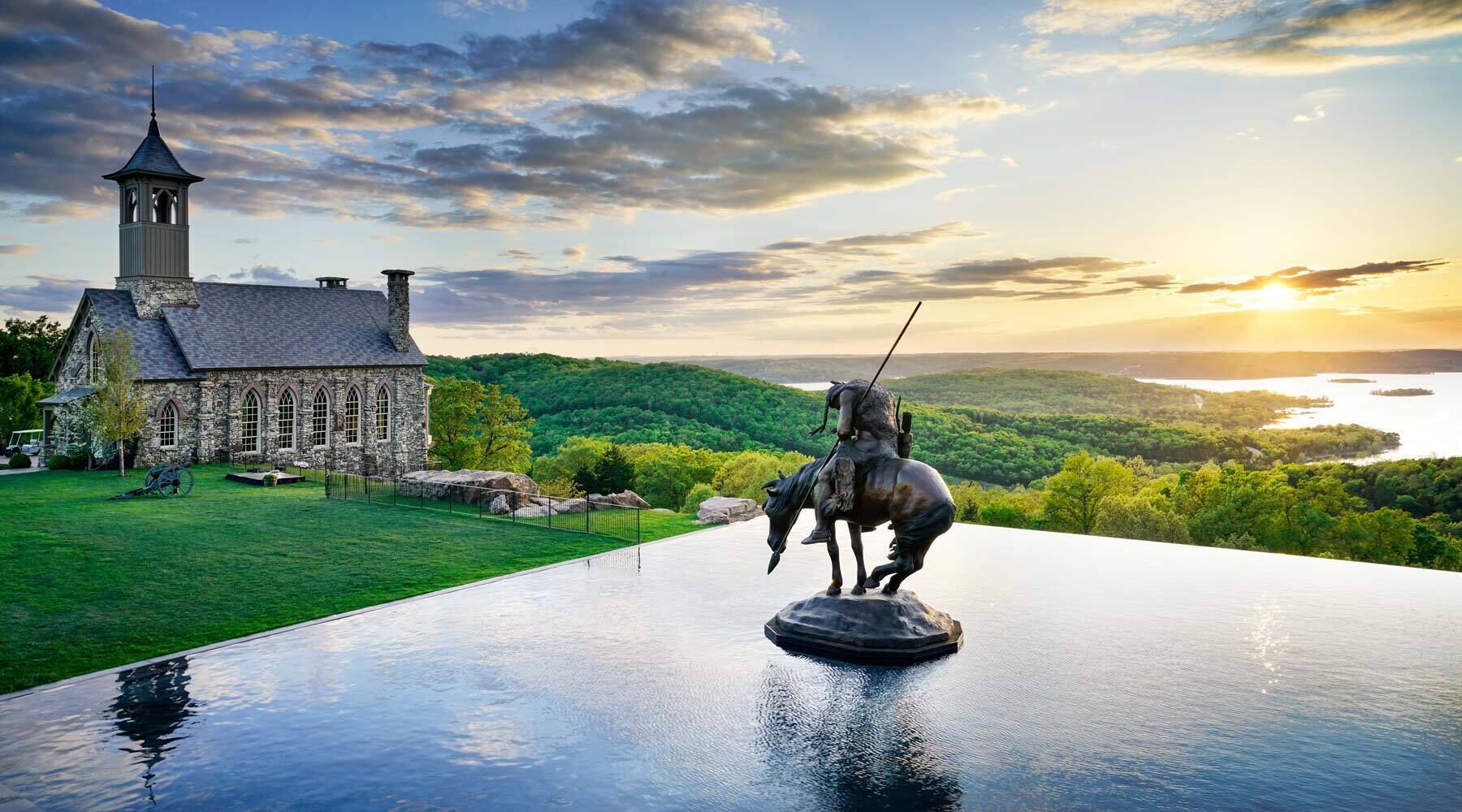 A stone church with a bell tower, a statue of a mounted figure, and a scenic view of a lake and forest.