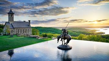 A stone church with a bell tower, a statue of a mounted figure, and a scenic view of a lake and forest.