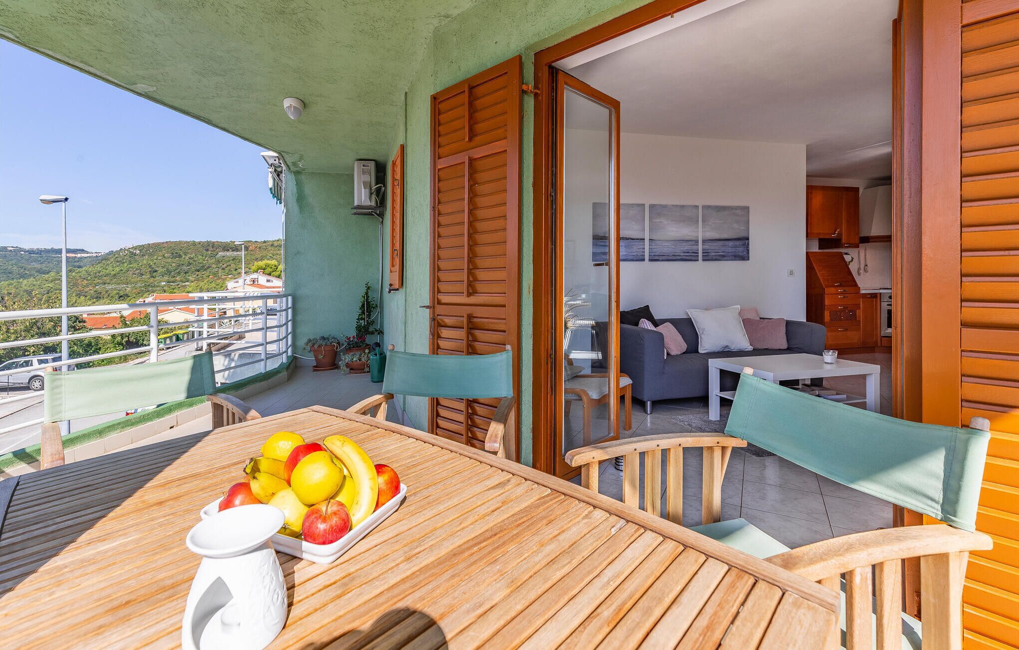 A wooden table with fruit, a sofa, and a dining area on a balcony with a view of the landscape.