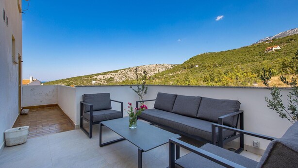 Een balkon met een bank, twee stoelen en een tafel. Het balkon kijkt uit op een heuvelachtig landschap met schaarse begroeiing.