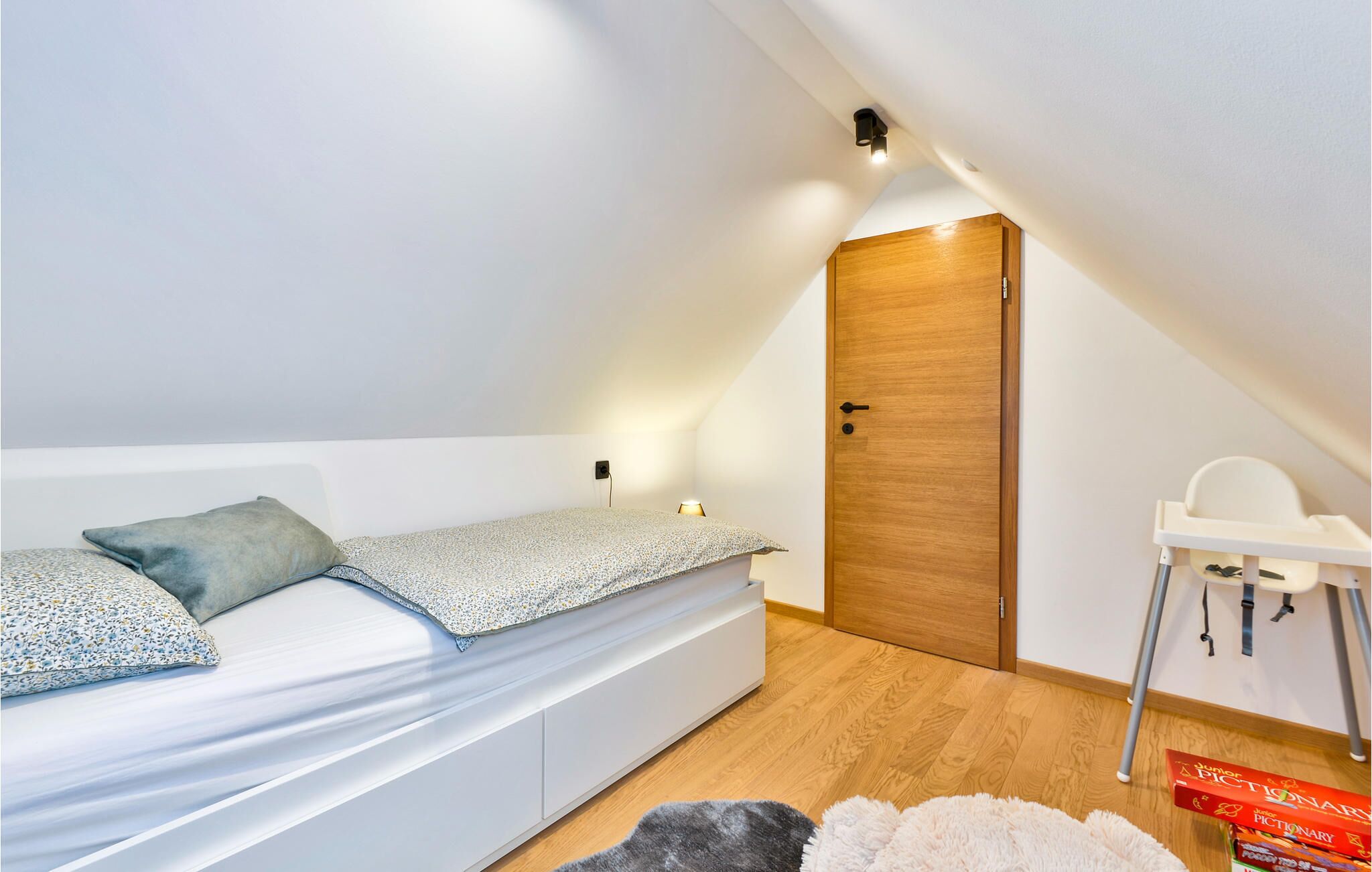 A small, well-lit attic bedroom with a single bed, a wooden door, and a white chair.