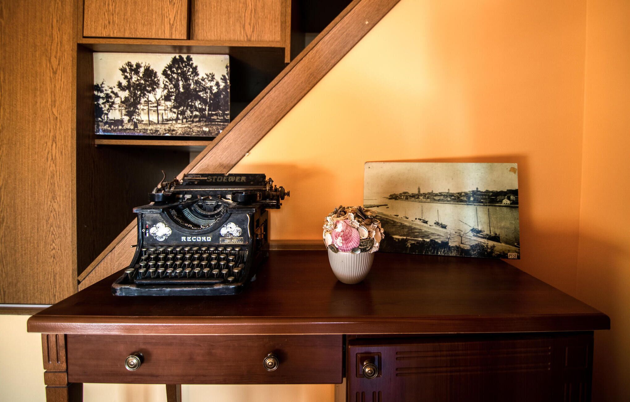 A vintage typewriter on a wooden desk with a vase of flowers and two framed photographs.