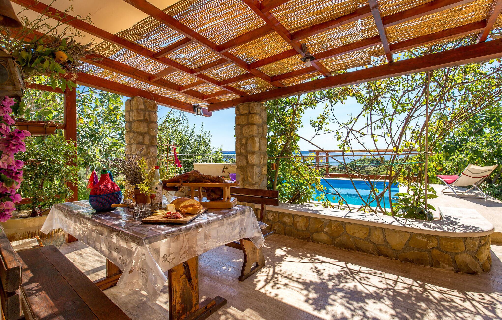 An outdoor dining area with a table set for a meal, a stone bench, and a view of a pool and mountains in the background.