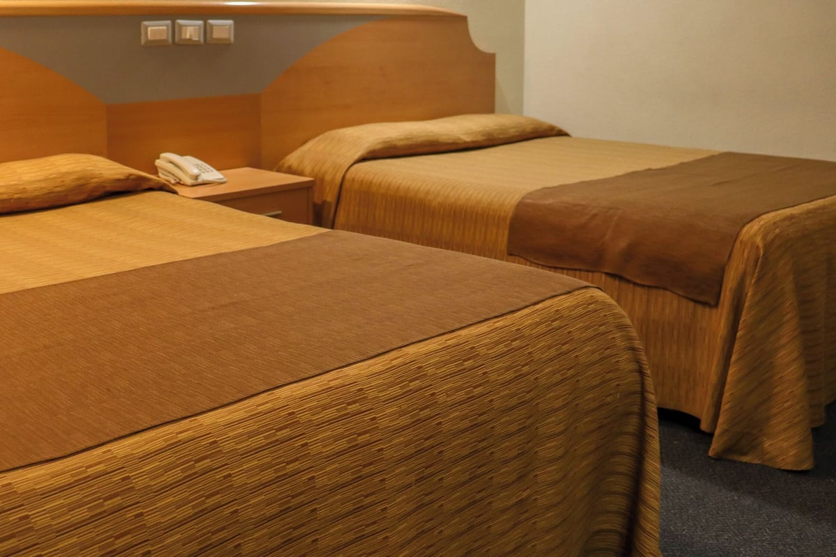 A hotel room with two beds, a wooden headboard, a bedside table with a telephone, and a wall-mounted light switch.