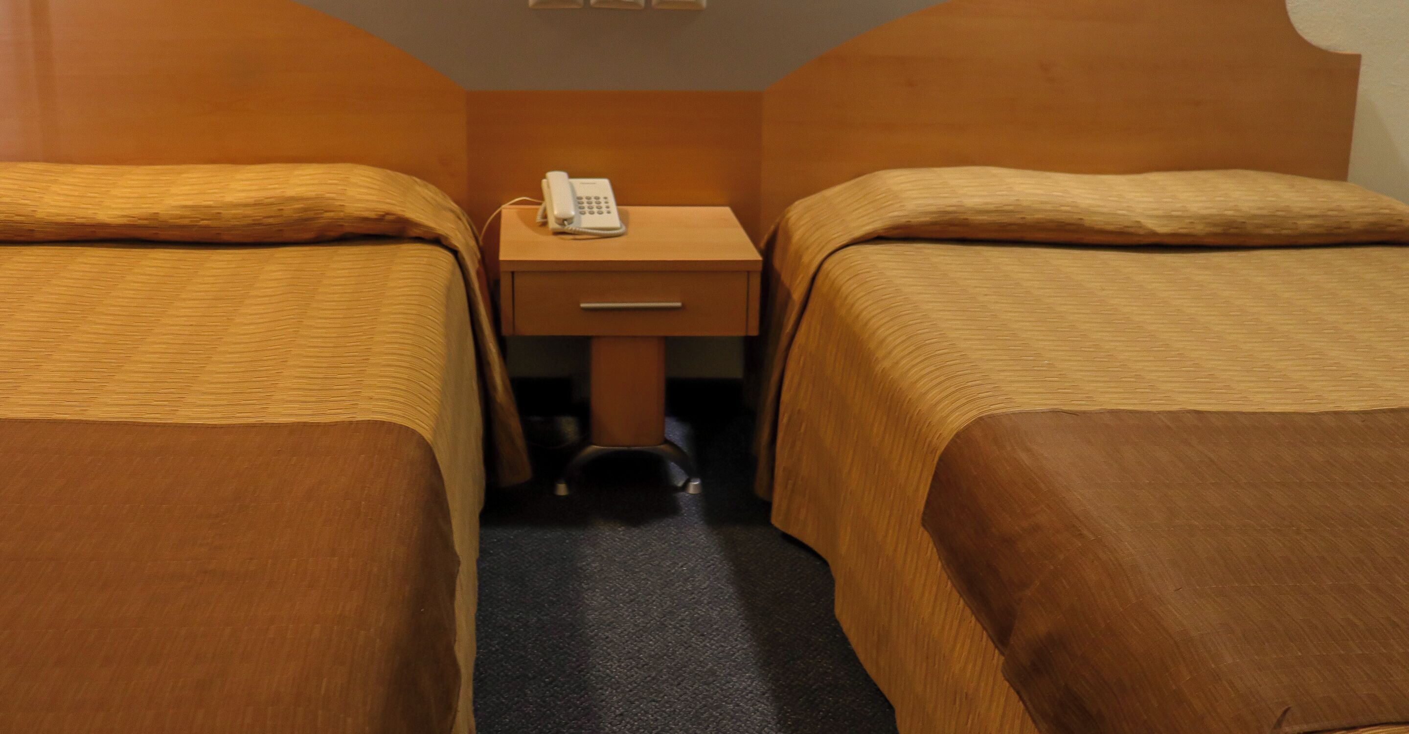 Una habitación de hotel con dos camas, una mesita de noche de madera con un teléfono, y piso alfombrado.