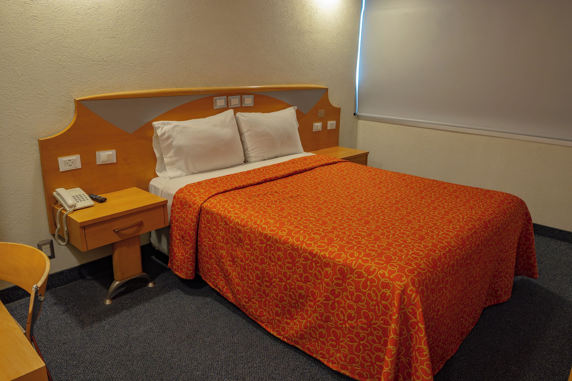 A hotel room with a bed, a bedside table with a phone, a chair, and a window.