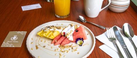 Un desayuno con un plato de fruta, yogur y granola, un vaso de zumo de naranja, una taza de café y un tazón pequeño de mantequilla y mermelada.