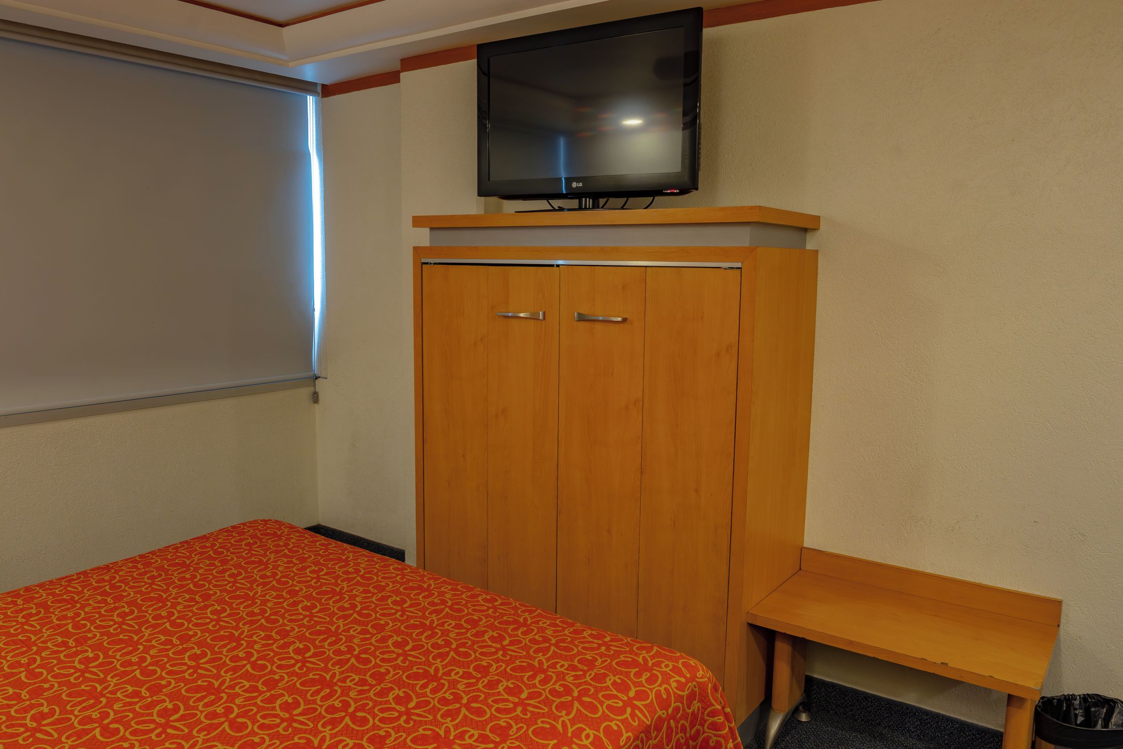 Una habitación con una cama, un armario de madera, un televisor encima y una ventana con persianas.