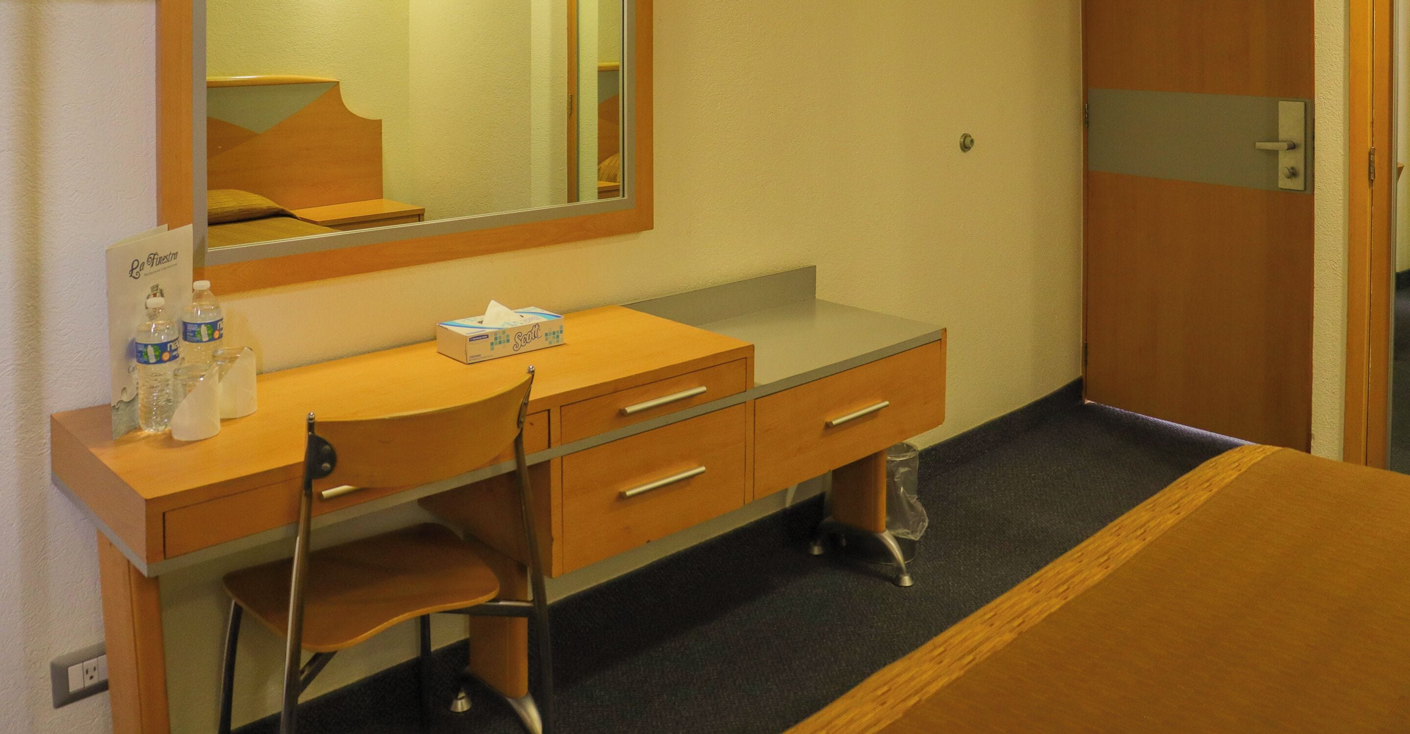 Ein Hotelzimmer mit Holzschreibtisch, Stuhl, Spiegel, Nachttisch und einer Tür.