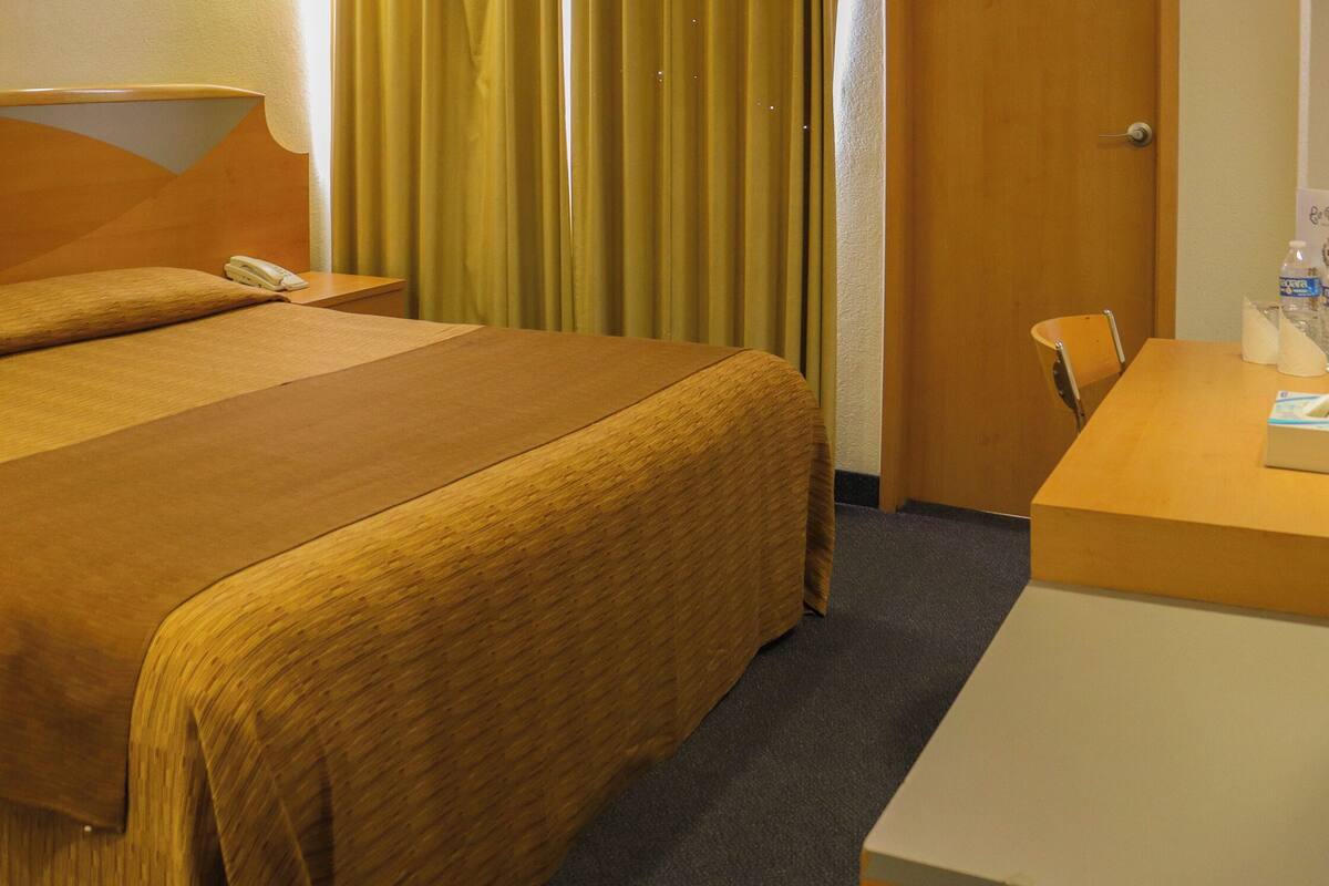A hotel room with a large bed, a bedside table with a lamp, a chair, and a desk with a bottle of water.