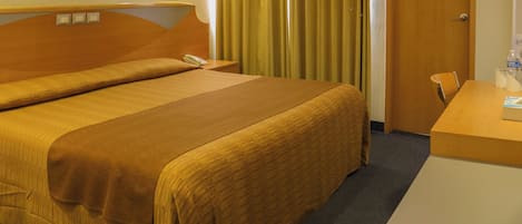 A hotel room with a large bed, a bedside table with a lamp, a chair, and a desk with a bottle of water.