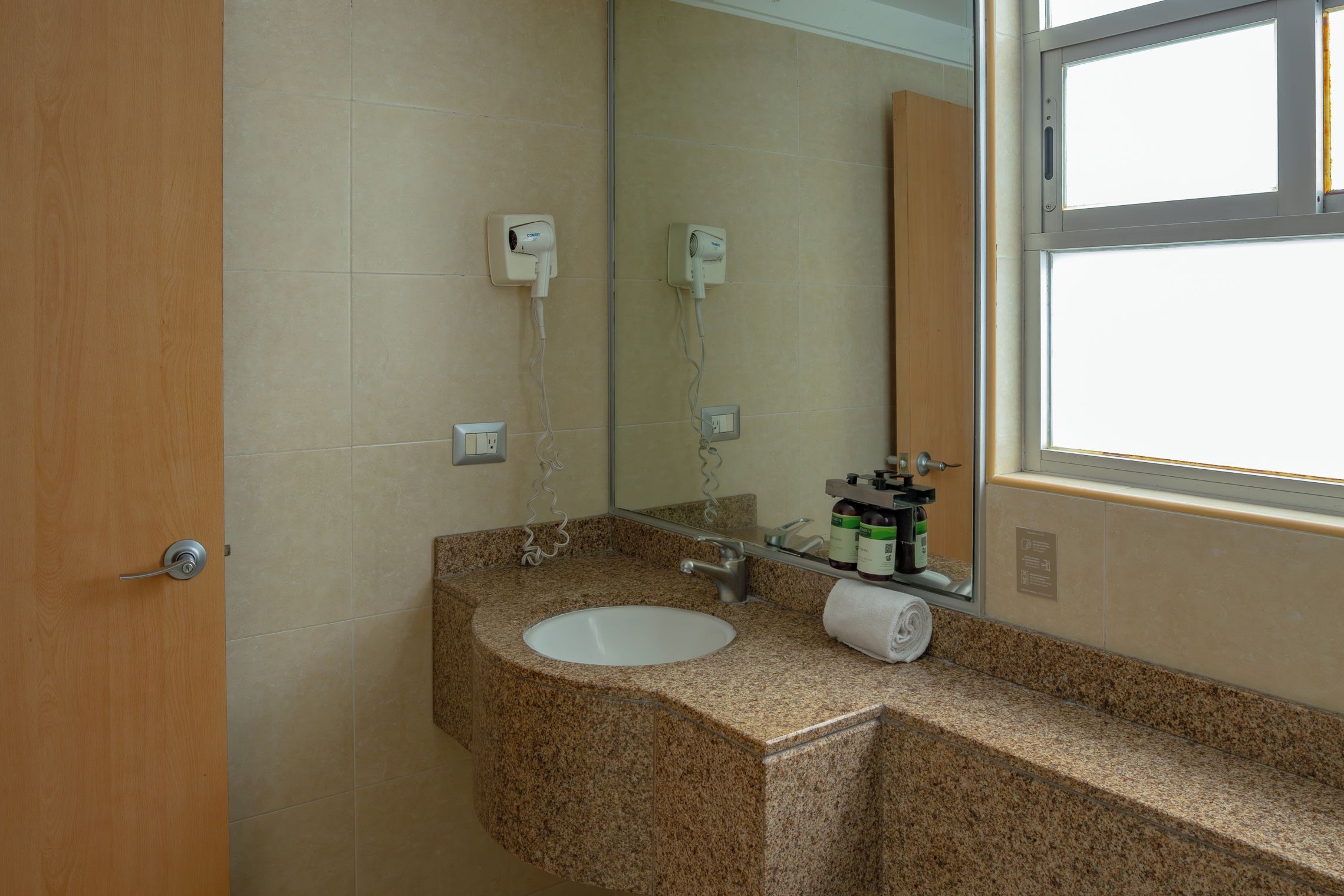 A bathroom with a granite countertop, a white sink, a mirror, and a hairdryer on the wall.