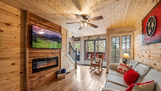 A wooden interior with a fireplace, a television mounted on the wall, and a ceiling fan. - Family Cabin Near Dollywood • Theater & Games (Pigeon Forge)