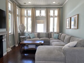 A living room with a sectional sofa, a fireplace, a TV, and large windows with curtains.