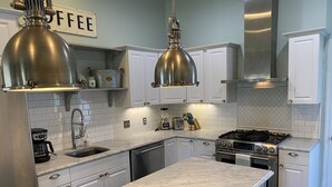 Cocina moderna con gabinetes blancos, encimera de mármol, electrodomésticos de acero inoxidable e iluminación colgante.
