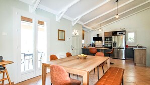 Un vaste espace de salle à manger avec une table en bois et des chaises rembourrées, une cuisine équipée d’appareils électroménagers modernes et une grande fenêtre laissant entrer la lumière naturelle.