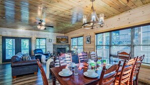 Un salon spacieux doté d’une cheminée, de grandes fenêtres et d’un coin repas avec des chaises en bois et une table dressée pour quatre personnes.