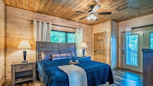 Une chambre à coucher dans une cabane en bois, comprenant un lit, deux tables de chevet, un ventilateur de plafond et une porte donnant sur une vue panoramique.