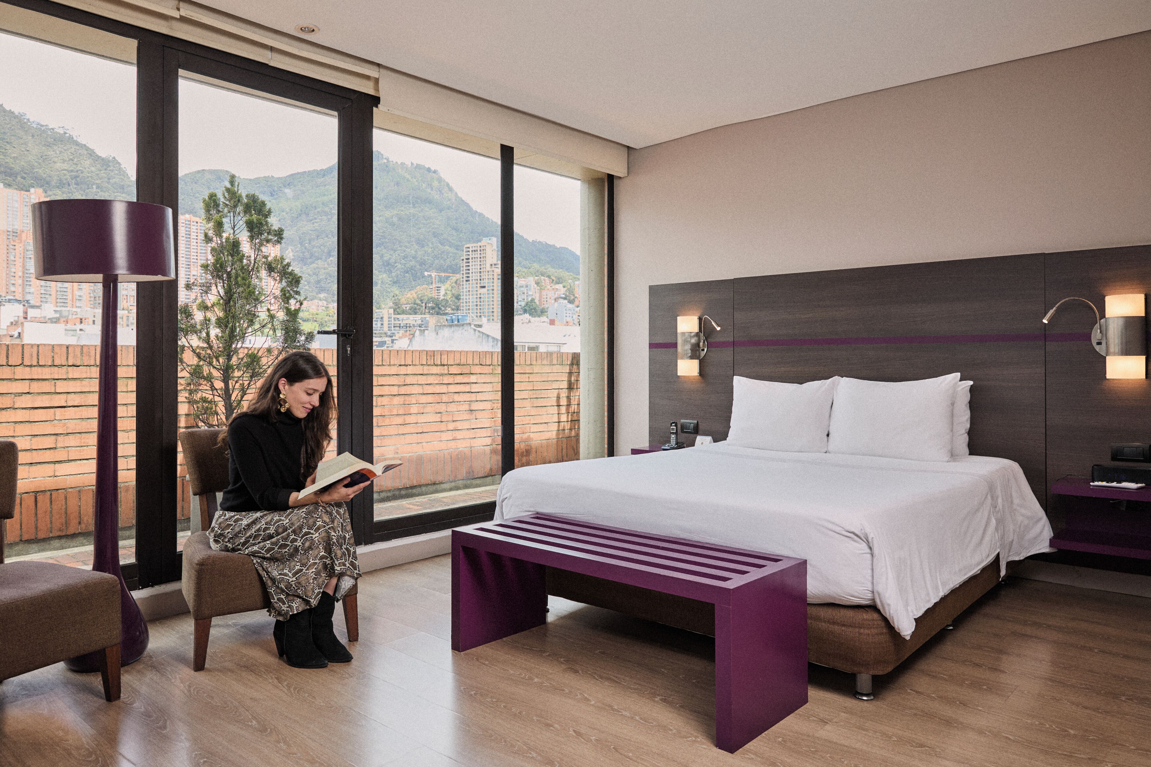 Habitación de hotel moderna con una cama grande, una banca para leer, un escritorio y vistas a un paisaje urbano a través de amplios ventanales.