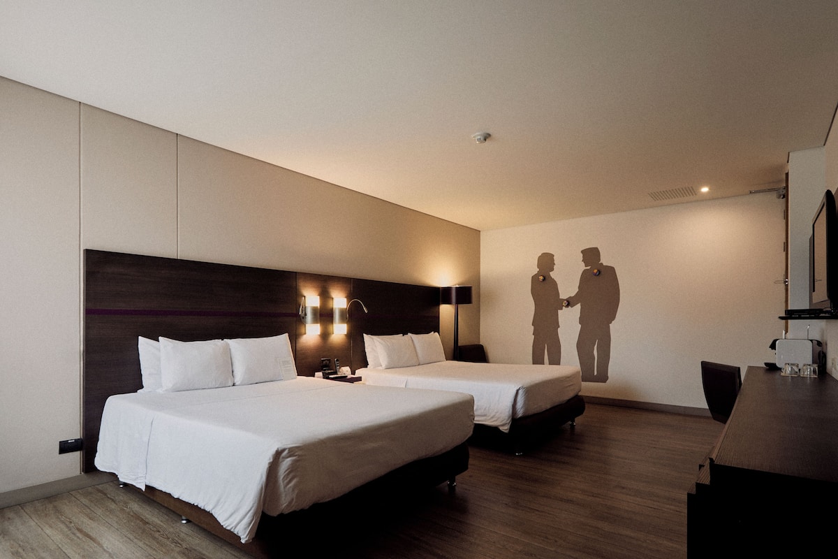 Habitación de hotel con dos camas, suelo de madera, un escritorio y un televisor montado en la pared.