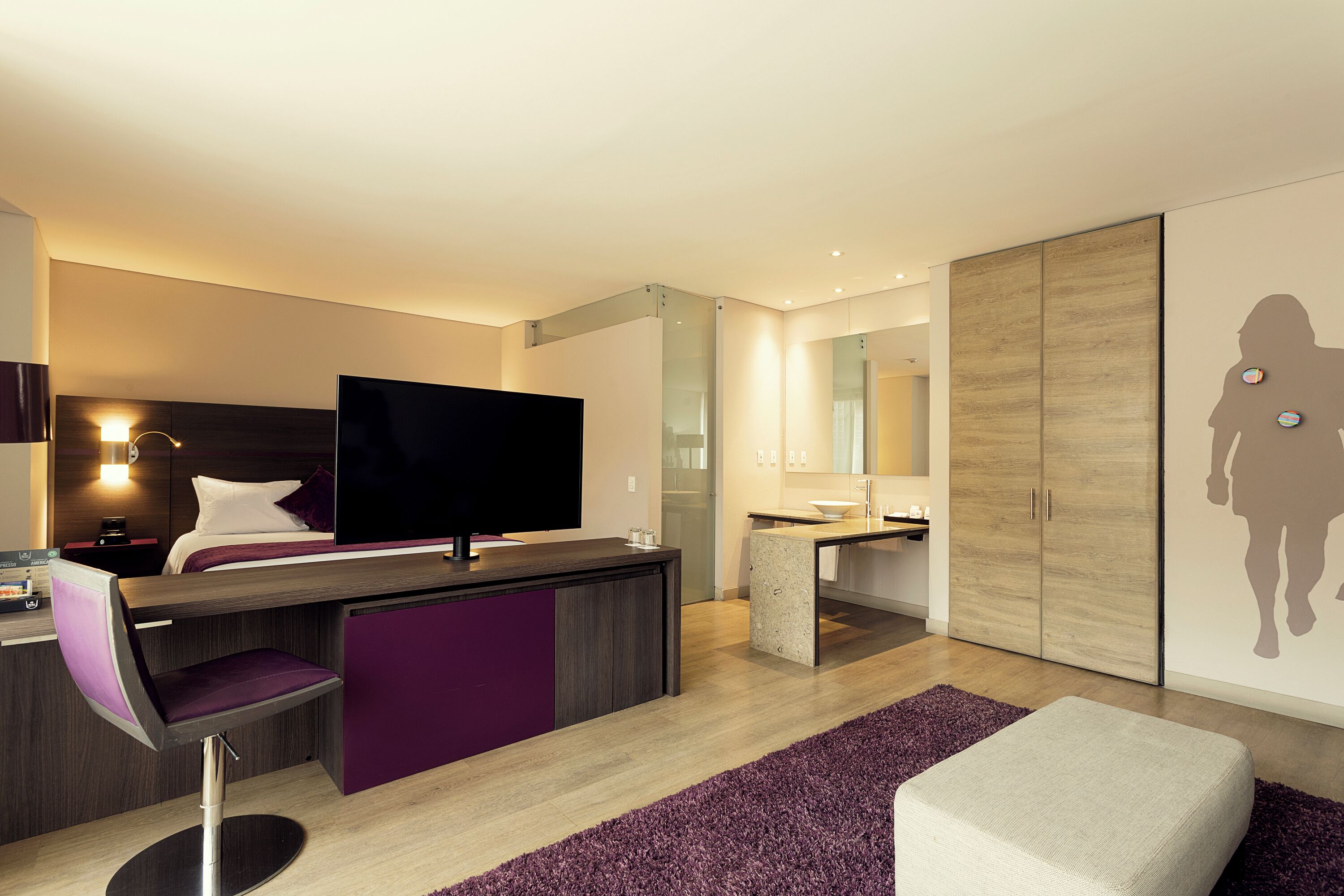 A modern hotel room with a flat-screen TV, a desk with a chair, a sofa, a large mirror, and a wardrobe.