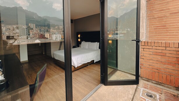 Habitación de hotel con cama, escritorio y vista a un paisaje urbano a través de una puerta de cristal.
