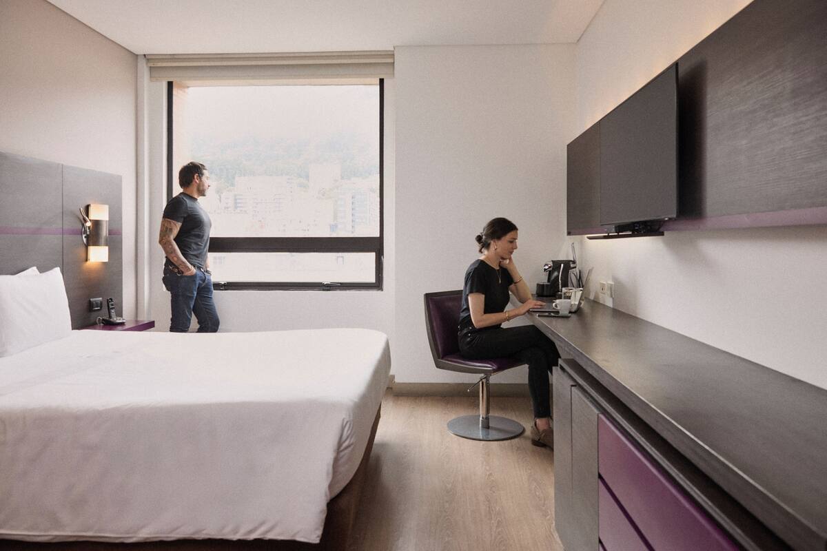 Habitación de hotel con cama, escritorio, silla, televisor de pantalla plana y ventana con vista a la ciudad.