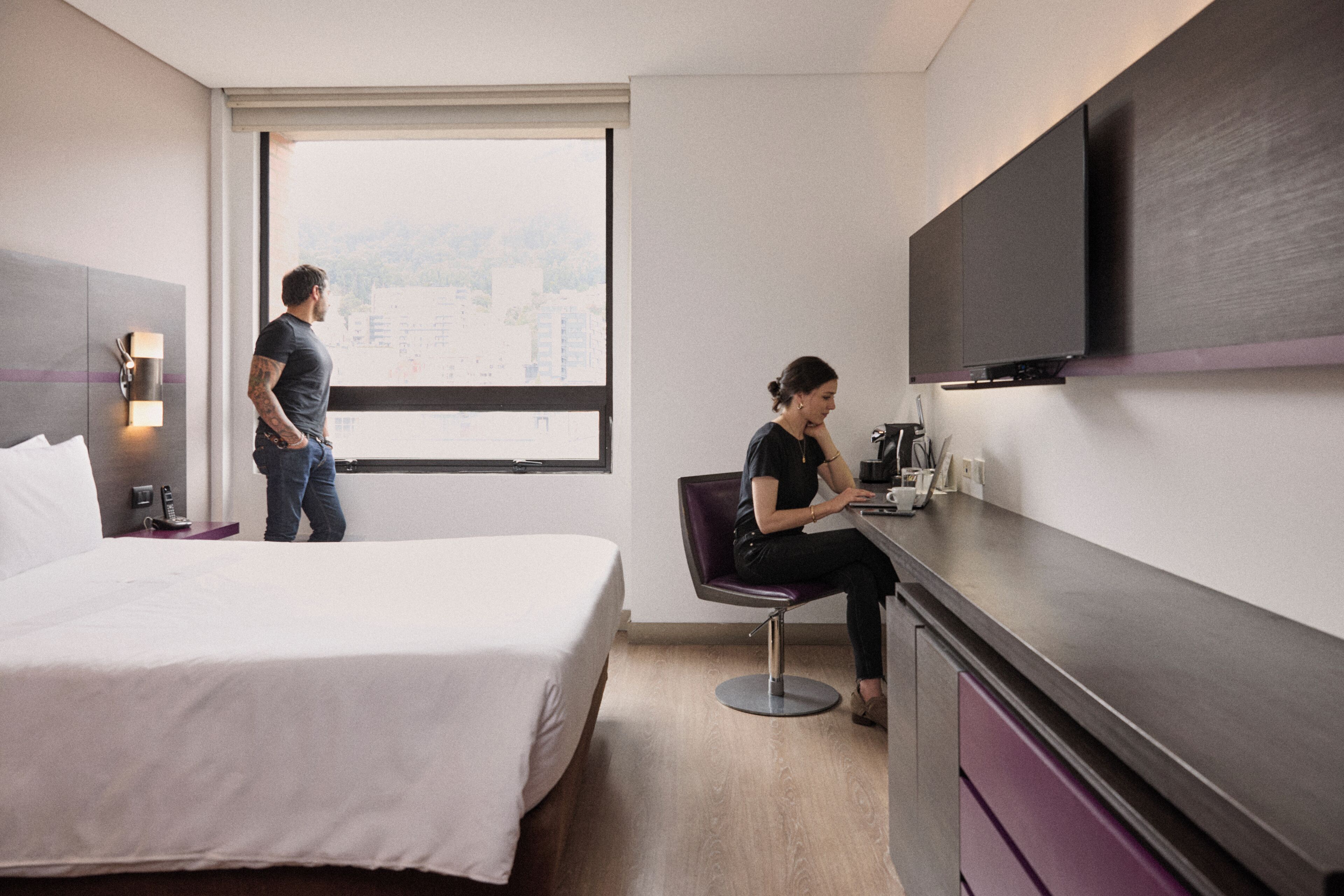 A hotel room with a bed, a desk, a chair, a TV, and a window with a city view.