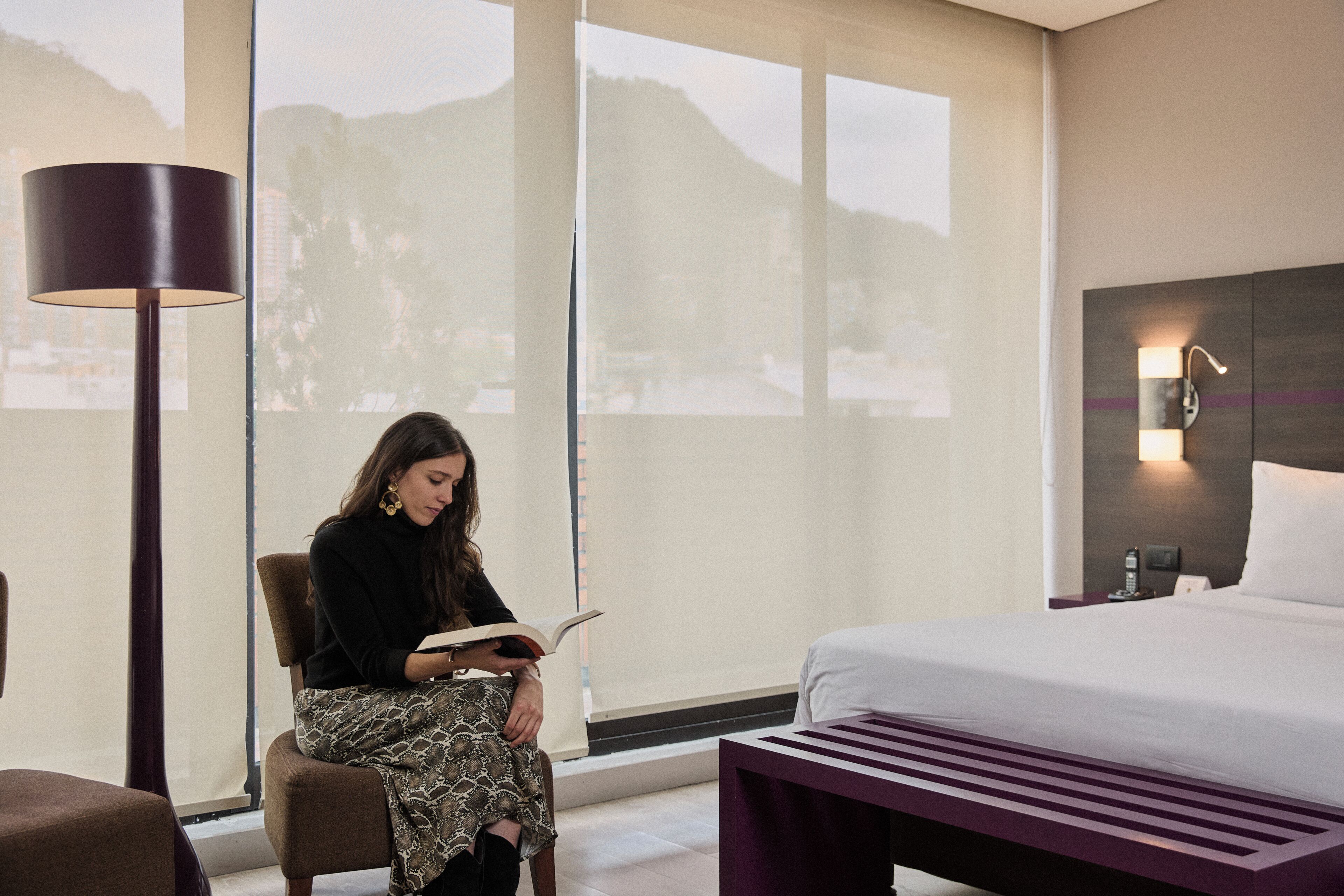 A hotel room with a bed, a reading chair, a large window with a view of mountains, and a wall-mounted lamp.