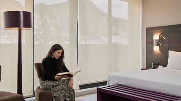 A hotel room with a bed, a reading chair, a large window with a view of mountains, and a wall-mounted lamp.