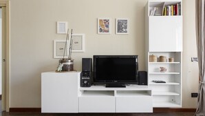 A living room with a TV on a white entertainment unit, framed pictures on the wall, and a bookshelf. - Hostmate - Milano - Teodorico (Milano)