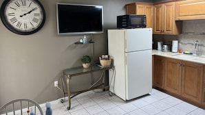 A kitchen with a wall-mounted clock, a flat-screen TV, a refrigerator, wooden cabinets, a tiled floor, and a small table with a plant. - Sunny Apartments a block from Warm Mineral Springs. Unit 3 (North Port)
