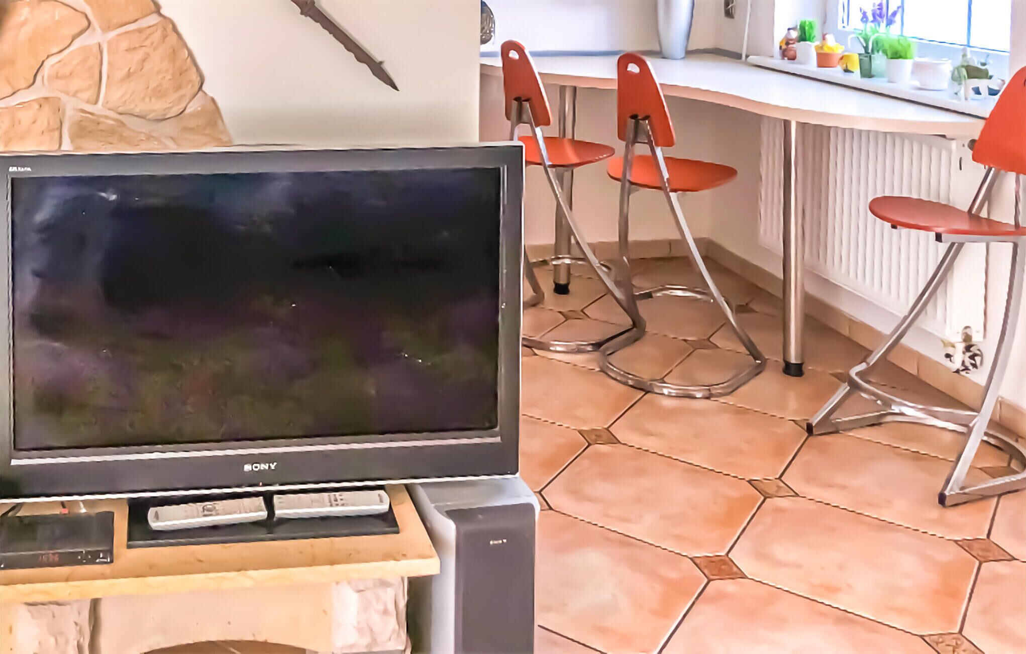A television on a wooden stand in a room with a stone wall, a tiled floor, and red chairs.