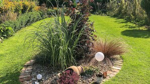Ein Garten mit verschiedenen Pflanzen, einem Ziegelweg und dekorativen Elementen wie einem weißen, kugelförmigen Objekt und einem Terrakottatopf.