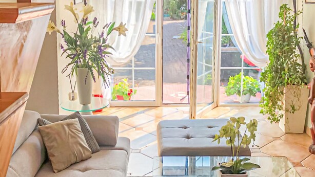 Ein modernes Wohnzimmer mit einer Couch, Hocker und einem Glastisch, mit Zimmerpflanzen und Blick durch große Glasfenster.