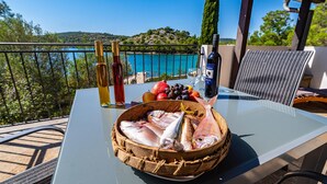 Ein Tisch mit einem Korb frischem Fisch, Weinflaschen und einem Glas Wasser auf einem Balkon mit Blick auf einen malerischen See.