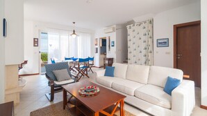 Un salon spacieux avec un canapé blanc, des fauteuils bleus, une table basse en bois et une vue sur la mer grâce à de grandes fenêtres.
