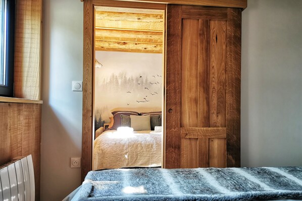 Un dormitorio con techo de madera, una cama con cubrecama blanco y vistas a un bosque a través de una puerta de cristal satinado.