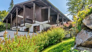 Um edifício em estilo chalé com fachada de madeira, várias varandas e um jardim com diversas plantas e um elemento decorativo de pedra.