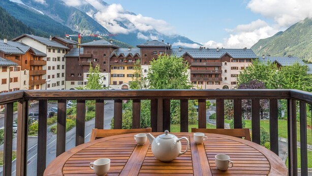 A wooden balcony with a table set for tea, overlooking a mountain village with traditional buildings and a snow-capped peak in the background.